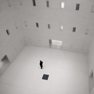 Black and white photo of a room with a very high ceiling, with three floors of windows very close to each other, several columns of windows. Three doors, one on each wall, visible in the photo. Only a man in black. And a square, also in black, in the middle of the room, on the floor. The man is close, located on the upper left side in relation to the square.